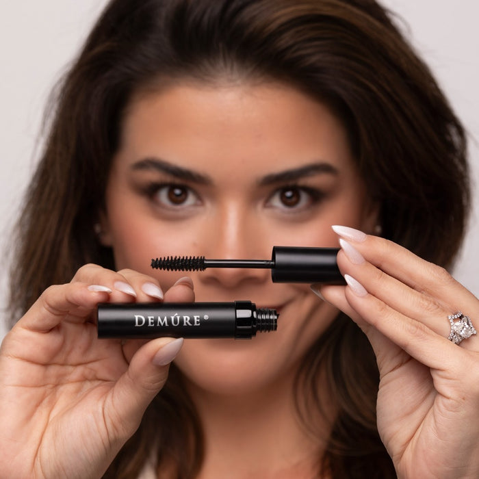 A close-up lifestyle shot of a woman holding a Demure Black Mascara tube and its wand, showcasing the product against her face.
