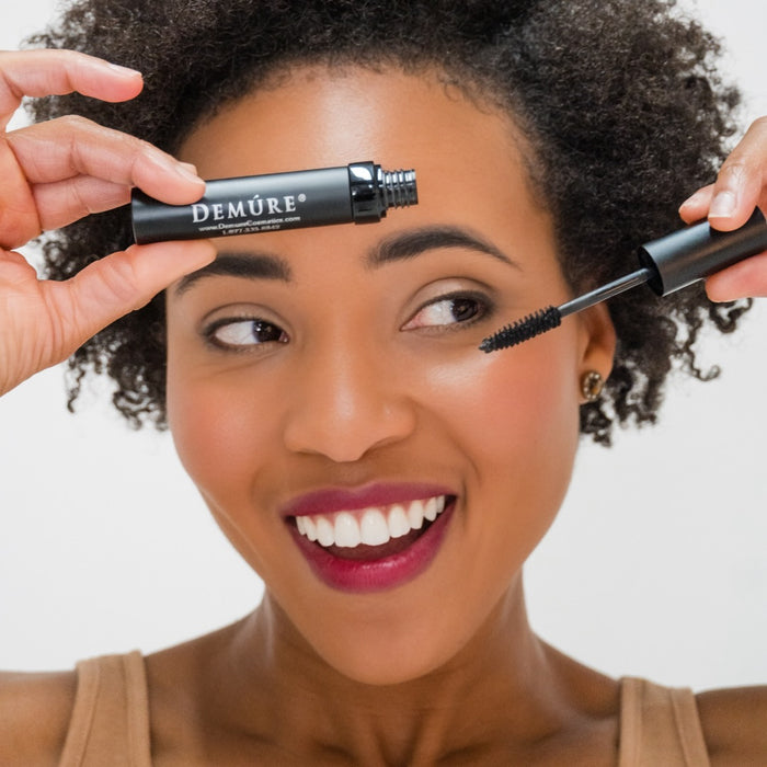 Woman applying mascara with a Demure mascara tube near her eye, against a neutral background