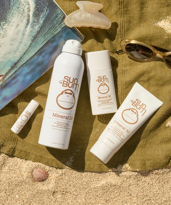 Sun Bum mineral sunscreen products on a sandy surface with a towel and sunglasses.