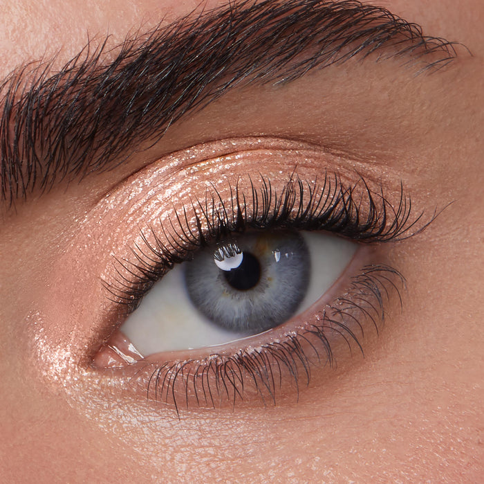Rose-gold shimmer eyeshadow gleams on a blue-gray eye, showcasing Mystical, a ColourPop Jelly Much Shadow, with wet-look metallic sheen and fine sparkle swept across lid and inner corner in a close-up beauty shot.