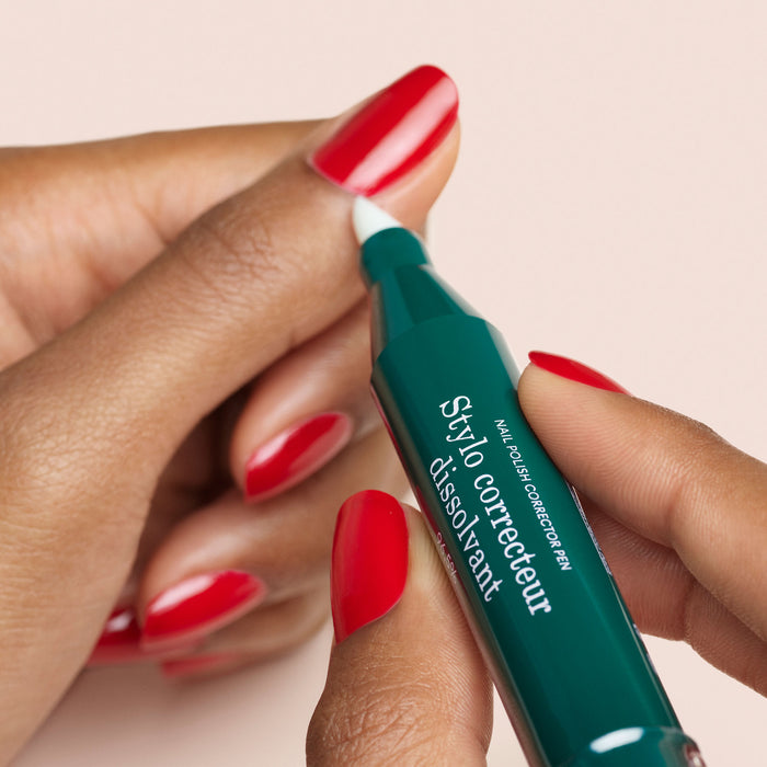 A person uses the Manucurist-test + Corrector Pen to clean up red gel polish on their fingernails, set against a light pink background.
