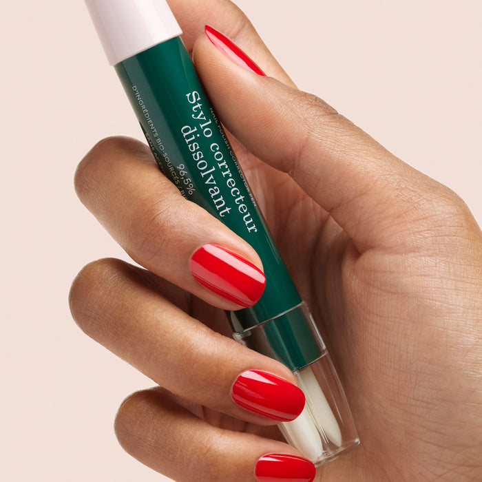 A hand with neatly manicured red nails holds the Manucurist-test + Corrector Pen, a green nail polish corrector pen, against a light beige background.
