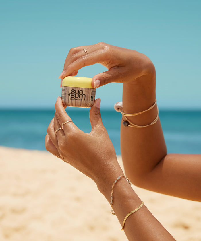 Hands holding a jar of Sun Bum Original SPF 50 Face Cream with a sandy beach and blue sky background