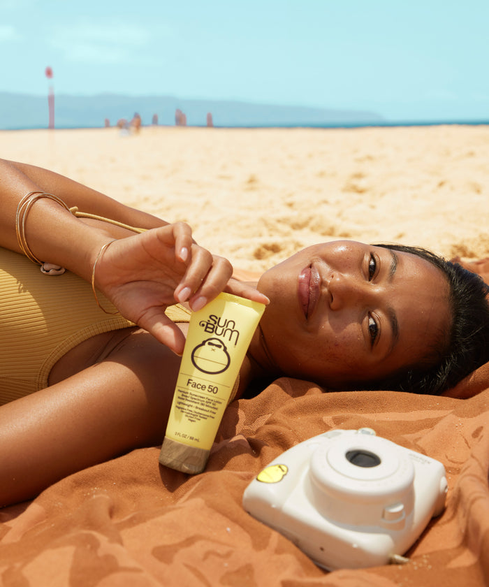 Woman relaxing on the beach holding Original SPF 50 Sunscreen Face Lotion by Sun Bum for lightweight sun protection.