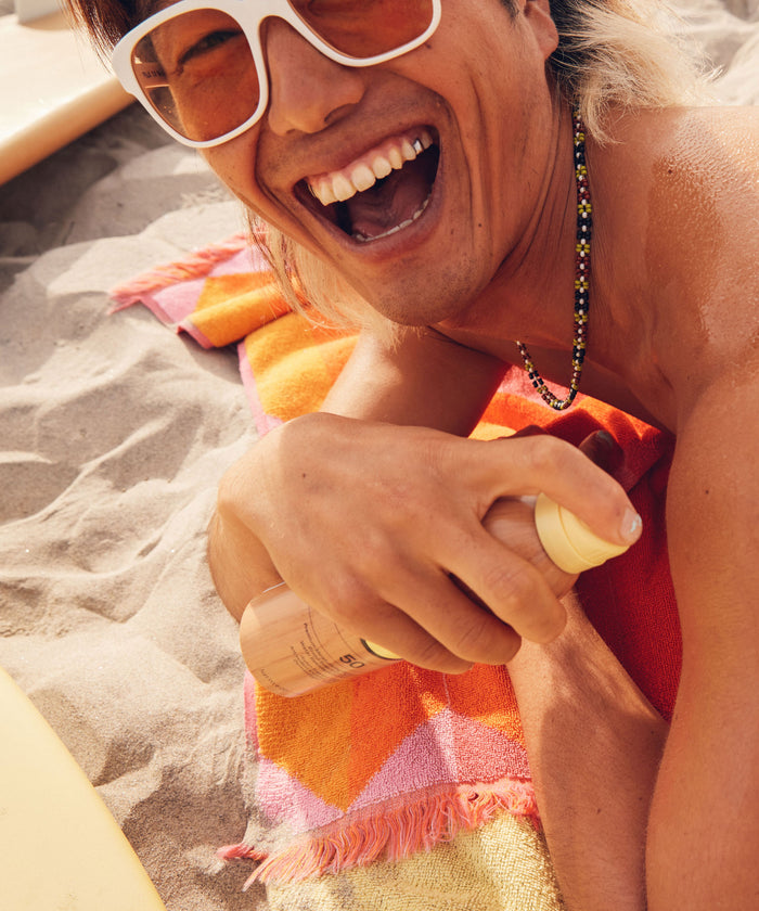 Person joyfully applying Original SPF 50 Sunscreen Spray on a beach, showcasing its ultra-sheer, dry touch formula that enriches skin with Vitamin E. Perfect for beach bags, using non-aerosol compression for application. Sun Bum.