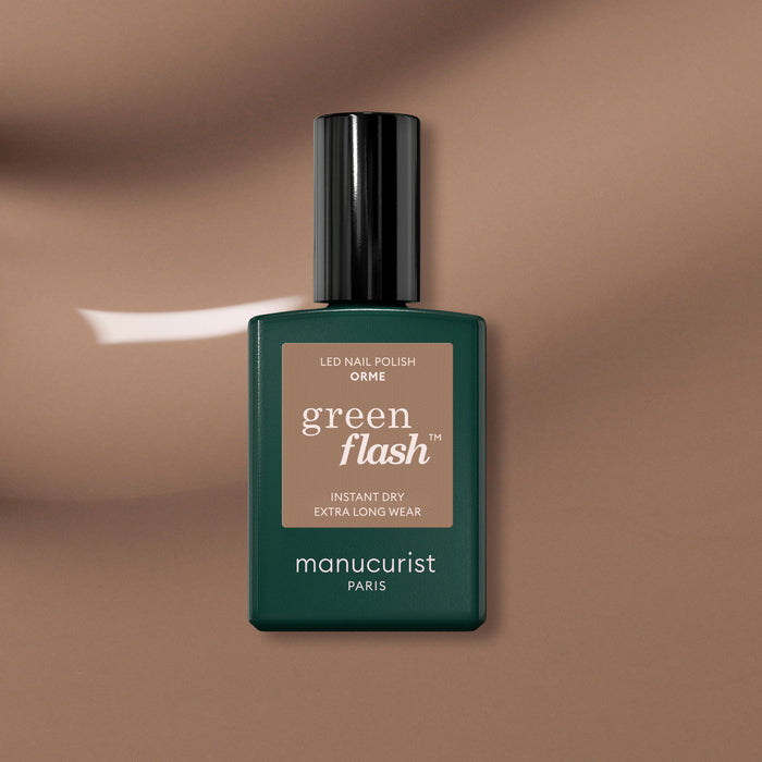 A bottle of Manucurist Orme gel polish, featuring a beige label and black cap, rests on a glossy brown surface. The Orme shade by Manucurist offers an eco-friendly update to classic gel polish.