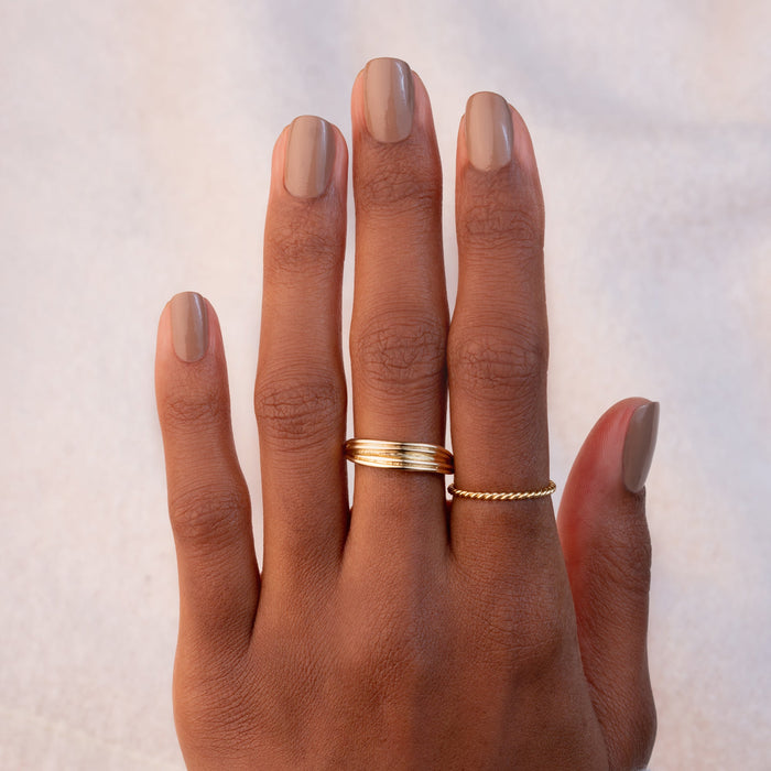 A hand with brown skin and neatly manicured nails painted in Manucurist's Orme polish, wearing a simple gold band on the middle finger and a twisted gold band on the ring finger, set against a soft, light background.