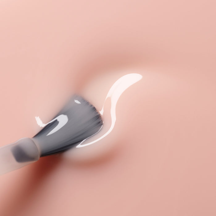 A close-up of a brush applying Manucurist's Pale Rose, a glossy translucent gel reminiscent of sheer pastel pink polish, onto a smooth peach-colored surface.
