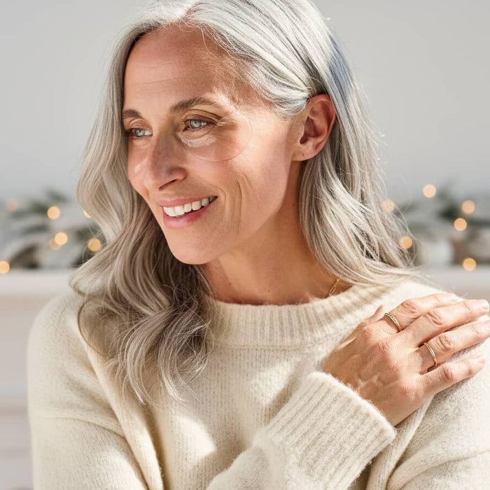 Woman with gray hair wearing a beige sweater, smiling with rose eye hydrating eye masks on.