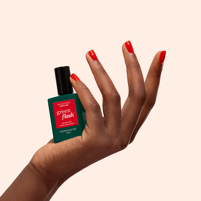A hand with Poppy Red-painted nails holds a bottle of Manucurist's Poppy Red polish against a light beige background.