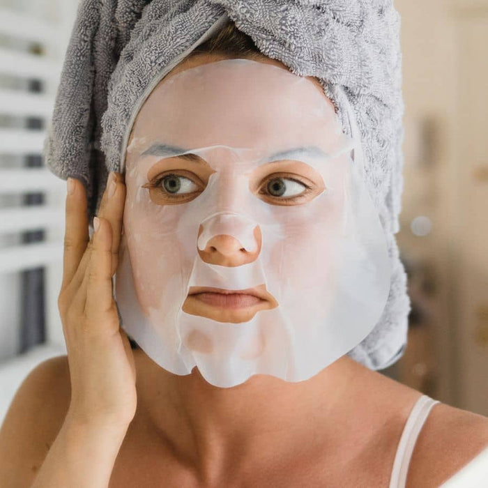 Woman wearing hydrating sheet face mask with towel-wrapped hair, gently touching cheek during skincare routine