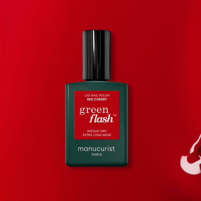 A bottle of Manucurist Red Cherry gel polish sits on a red background. The dark green bottle features a black cap and displays the product name on a red label.