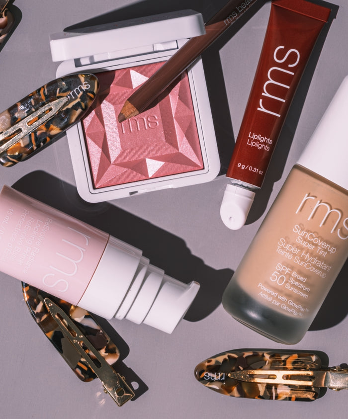 Flat lay of the RMS Discovery Kit featuring skincare infused makeup including a pink blush, tinted lip oil, sunscreen with SPF 50, a pencil, and accessories on a gray surface.