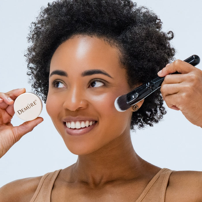 African American woman wearing Demure Pink Fusion mineral blush with a contour brush and holding a jar against a neutral