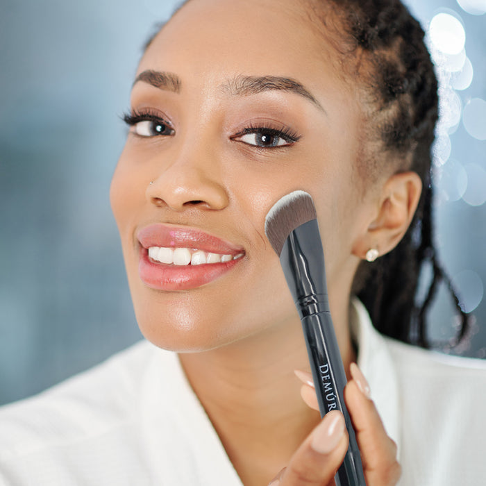 A woman holding a Sculpting Contour Brush next to her right cheek.