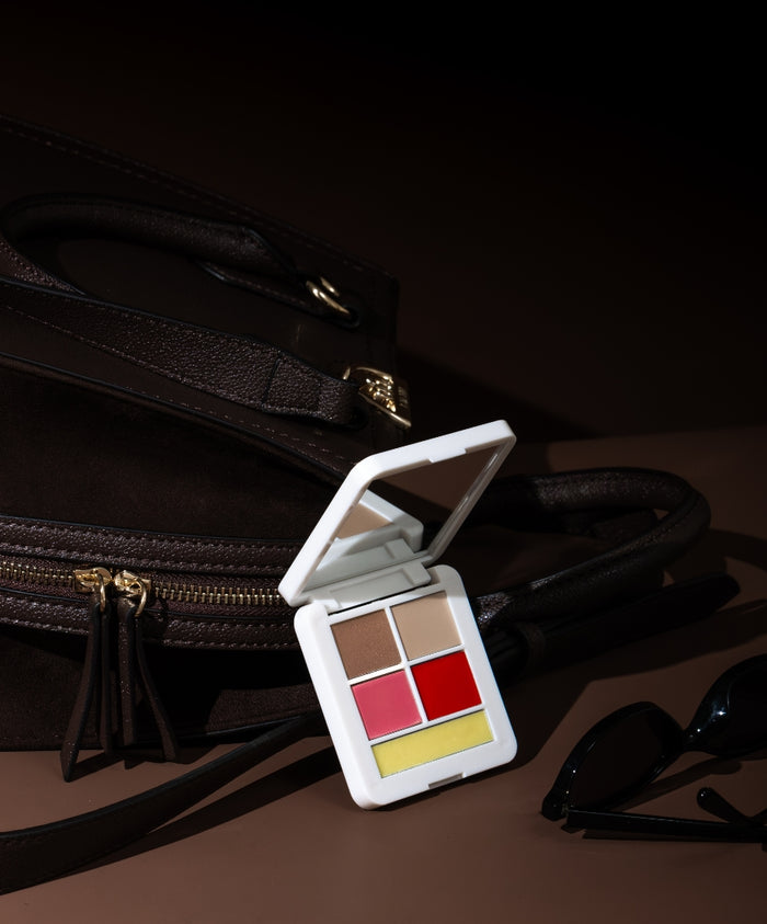 Compact white makeup palette featuring neutral and bold shades with a mirror, set against a dark brown leather bag and black sunglasses, showcasing the Signature Set.