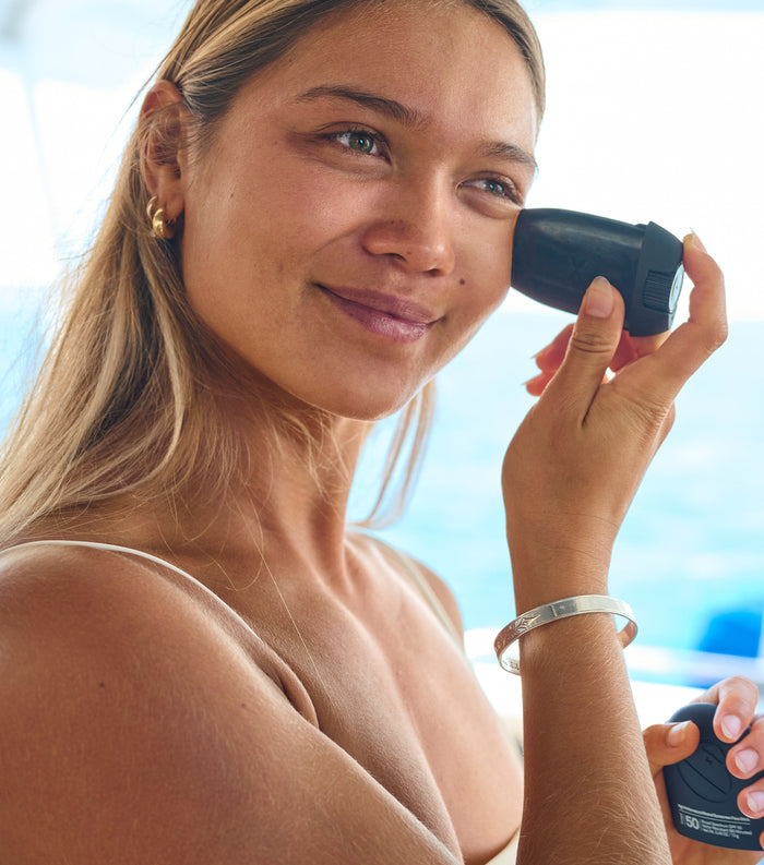 Woman applying Sun Bum Signature SPF 50 Tinted Mineral Face Stick to her cheek with a smile outdoors