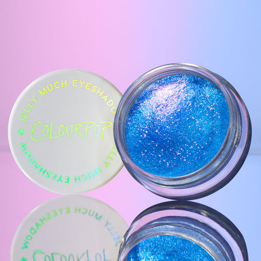 ColourPop Jelly Much Shadow eyeshadow in Stargaze open jar showcasing shimmering electric blue jelly with pink-violet sparkles, the branded white lid beside it on a mirrored surface against a pink-to-blue gradient backdrop.
