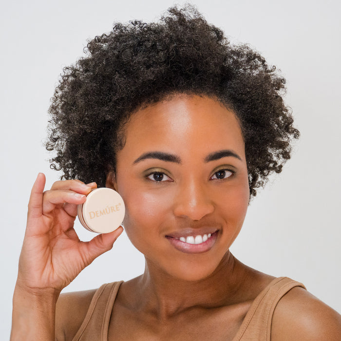 African American woman wearing Deluvia Dark Warm Foundation and holding the jar near her cheek.