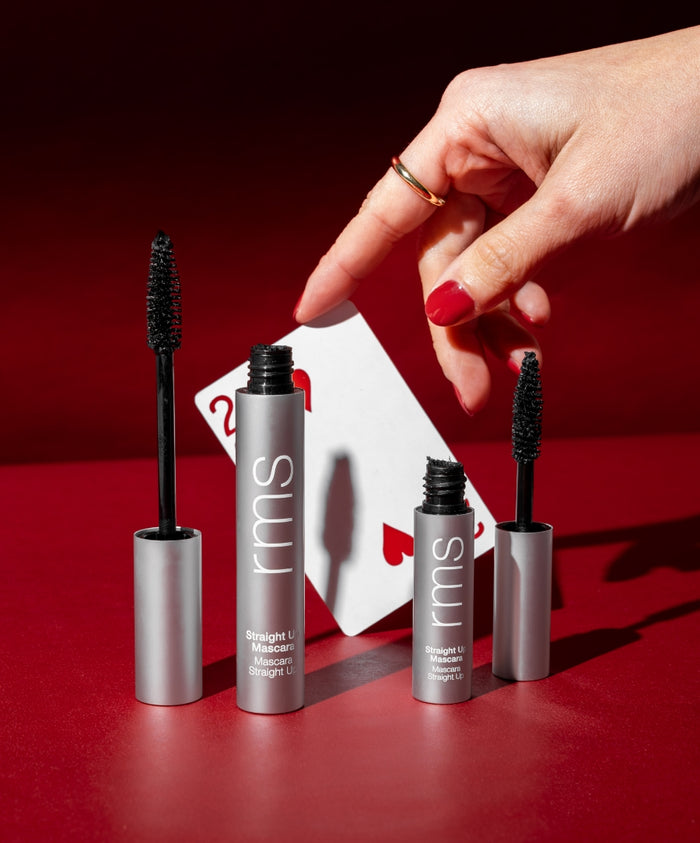 Hand with red nail polish and gold ring holding a playing card behind two open tubes of Straight Up™ Volumizing Peptide Mascara with black brush wands on a red background, showcasing clean beauty and sustainable packaging.