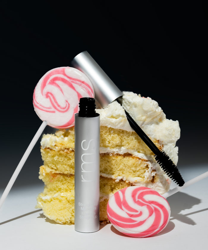 Straight Up™ Volumizing Peptide Mascara with black brush wand displayed alongside slices of vanilla cake and pink-and-white swirl lollipops on a dark background, showcasing clean beauty and natural ingredients.