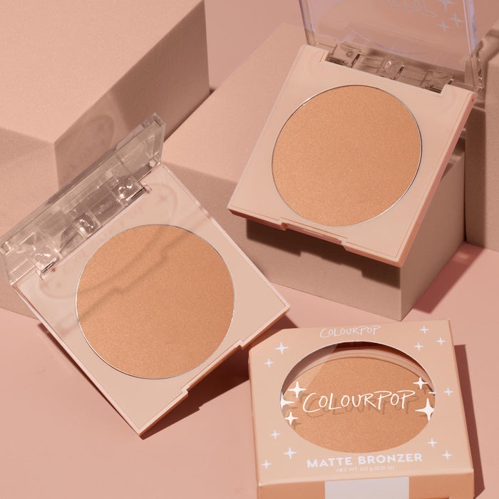 ColourPop Summerland Beach pressed powder bronzer compact sits open to reveal a warm matte tan pan, alongside a second open compact and a boxed unit on blush‑pink blocks; ColourPop Pressed Powder Bronzer for a natural sun‑kissed finish.