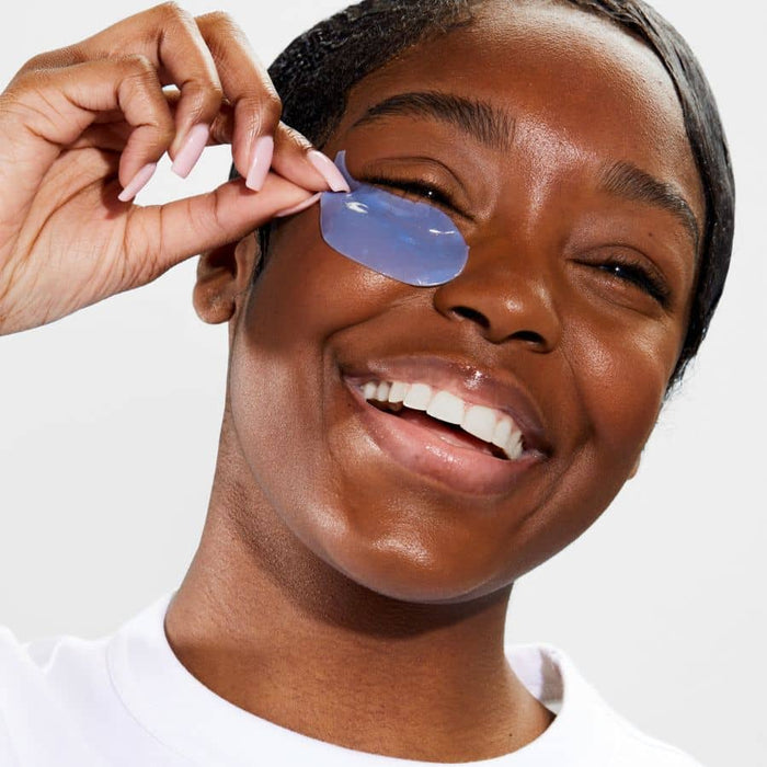Smiling woman with glowing skin applying a blue hydrogel eye patch for a cooling and depuffing skincare routine.
