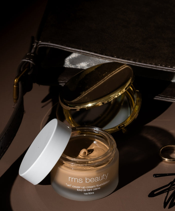 Open jar of UnCoverup Cream Foundation by RMS Beauty with creamy beige foundation inside, placed next to a compact mirror and dark leather bag on a brown surface.