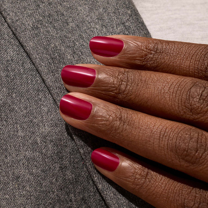 A close-up of a hand with short, neatly manicured nails painted in Manucurist's Violeta polish, resting on gray textured fabric.