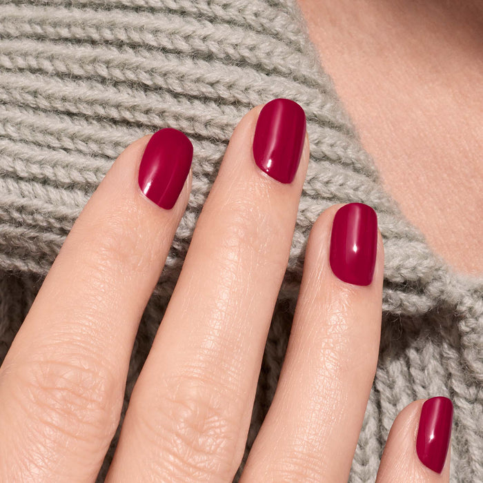 A hand with neatly manicured nails painted in Manucurist's Violeta, a glossy burgundy purple polish, rests on textured light gray knit fabric.