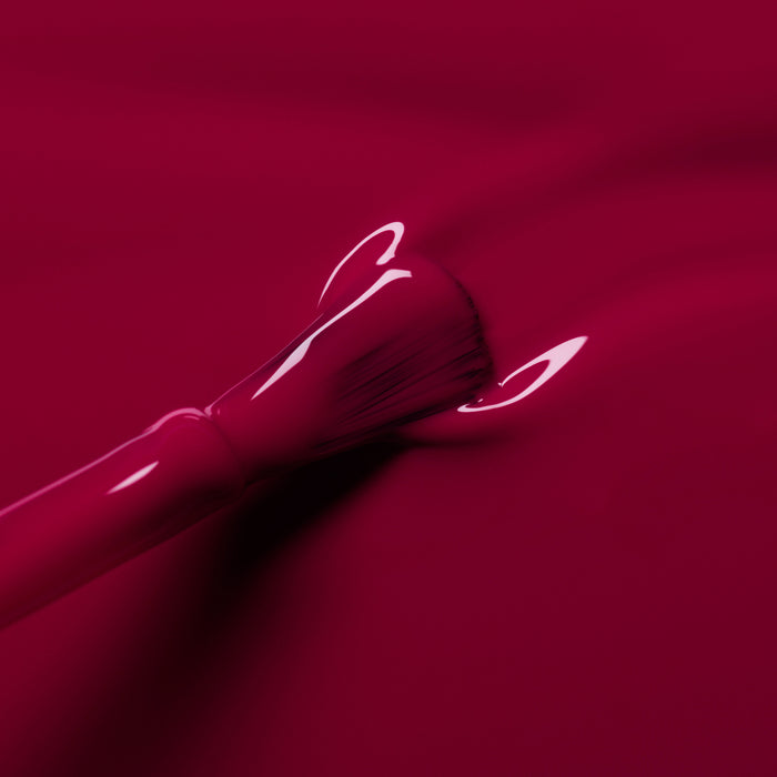 A close-up of a nail polish brush spreading Manucurist's Violeta, a glossy burgundy purple polish, across a smooth surface.
